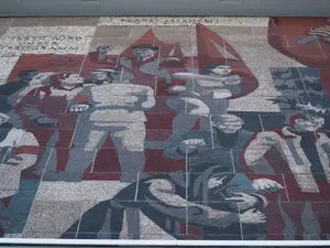 KI generiert: Das Bild zeigt ein großes Wandbild mit stilisierten, abstrakten Figuren, die in einer Mischung aus Rot-, Grau- und Blautönen dargestellt sind. Es scheint eine historische oder politische Szene darzustellen, die möglicherweise Arbeiter oder revolutionäre Figuren zeigt.