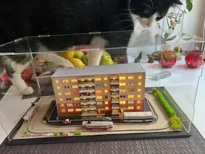 KI generiert: Das Bild zeigt ein Miniatur-Modell eines Gebäudekomplexes mit beleuchteten Fenstern und kleinen Fahrzeugen auf einer Straße. Eine Katze steht vor dem Modell und zeigt Interesse daran.
