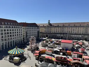 KI generiert: Das Bild zeigt einen belebten Platz mit einem Jahrmarkt, auf dem es zahlreiche Buden, ein kleines Karussell und ein Riesenrad gibt. Umgeben ist der Platz von großen, mehrstöckigen Gebäuden in einem urbanen Umfeld.