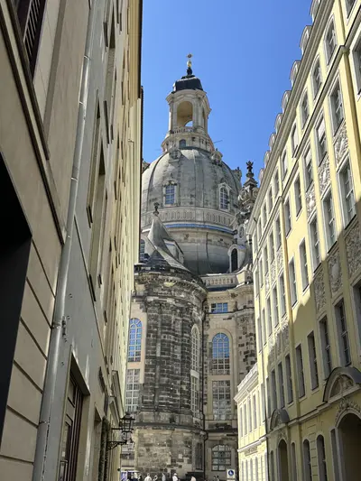 KI generiert: Das Bild zeigt die Frauenkirche in Dresden, gesehen aus einer engen Gasse zwischen Gebäuden. Der markante Kuppelbau ist der zentrale Fokus des Bildes.