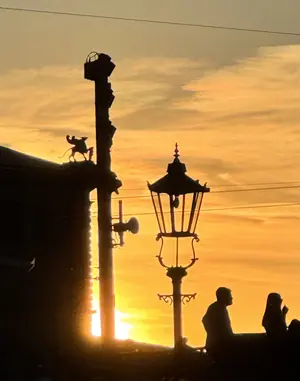 KI generiert: Das Bild zeigt eine Silhouette-Szene bei Sonnenuntergang, mit einer Straßenlaterne und Personen auf einer Brücke. Der Himmel leuchtet in warmen Gelb- und Orangetönen.