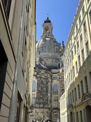 KI generiert: Das Bild zeigt die Frauenkirche in Dresden, gesehen aus einer engen Gasse zwischen Gebäuden. Der markante Kuppelbau ist der zentrale Fokus des Bildes.